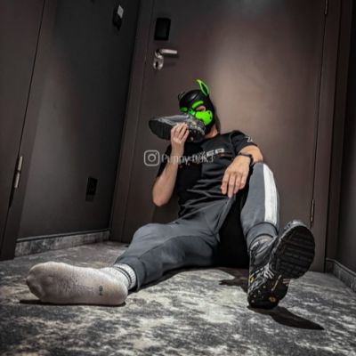 Person wearing a black and green puppy play hood, black sportswear and grey socks, sitting on the floor while holding a black sneaker. The sporty puppy play outfit and well-worn socks create a football-inspired fetish aesthetic with a focus on sneakers, socks and athletic gear.