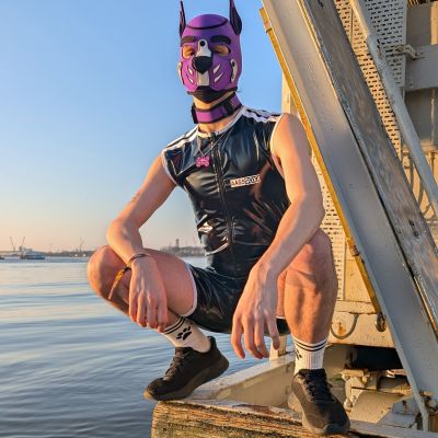 Person wearing a purple puppy play hood, black shiny PVC singlet, white crew socks and black sports sneakers, crouching by the waterfront at sunset. The glossy PVC outfit and athletic styling create a striking sports fetish look with puppy play gear in an industrial outdoor setting.