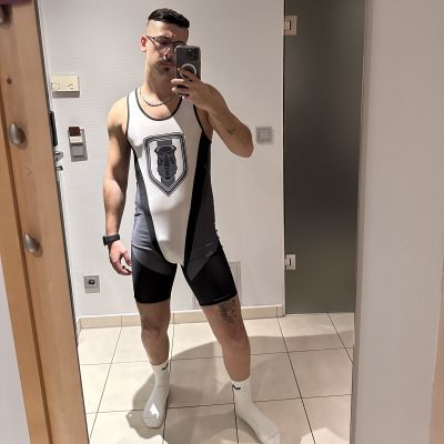 Man taking a mirror selfie wearing a black, white and grey wrestling singlet with a bulldog emblem and white Nike crew socks. The fitted athletic gear highlights a muscular physique, creating a clean sportswear aesthetic with wrestling and men's fetish fashion influences.