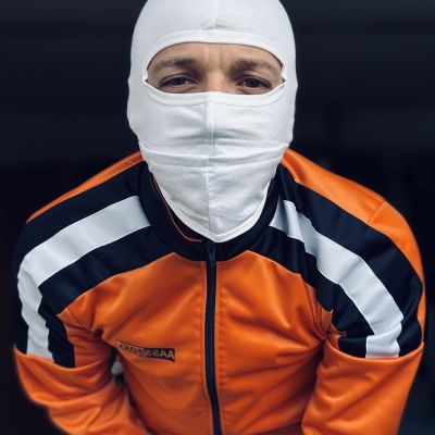 Person wearing an orange, black and white ASOX tracksuit jacket with a white balaclava, posing for a close-up portrait. The sporty outfit combines technical sportswear with a bold fetish-inspired aesthetic focused on athletic fashion and face covering.