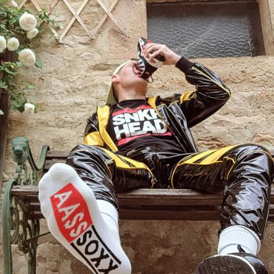 Person wearing a black and yellow shiny PVC tracksuit, white ASOX crew socks and black sports sneakers while smelling a trainer outdoors. The glossy sportswear, branded socks and sneaker-focused pose create a bold sneaker fetish aesthetic with urban sports style.