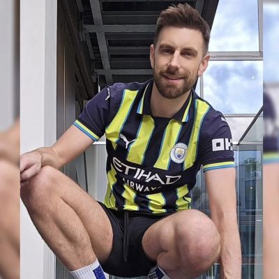 Smiling man wearing a Manchester City football jersey, black athletic shorts and white crew socks, crouching outdoors on a modern balcony. The sporty outfit and confident pose create a football-inspired, sportswear aesthetic with an urban background.