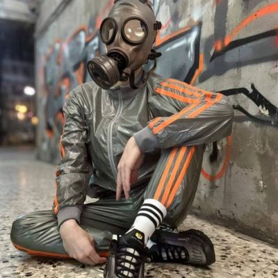 Person wearing a vintage gas mask, grey shiny tracksuit with orange stripes, white striped crew socks and black sports sneakers, sitting against a graffiti-covered wall. The industrial-inspired sportswear creates a striking urban fetish aesthetic featuring technical fabrics, sneakers and athletic styling.