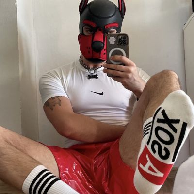 Person wearing a red and black puppy play hood with a matching collar, white compression shirt, shiny red PVC shorts and white ASOX crew socks, taking a mirror selfie. The sporty puppy play outfit combines athletic gear, PVC clothing and branded socks for a bold fetish-inspired look.